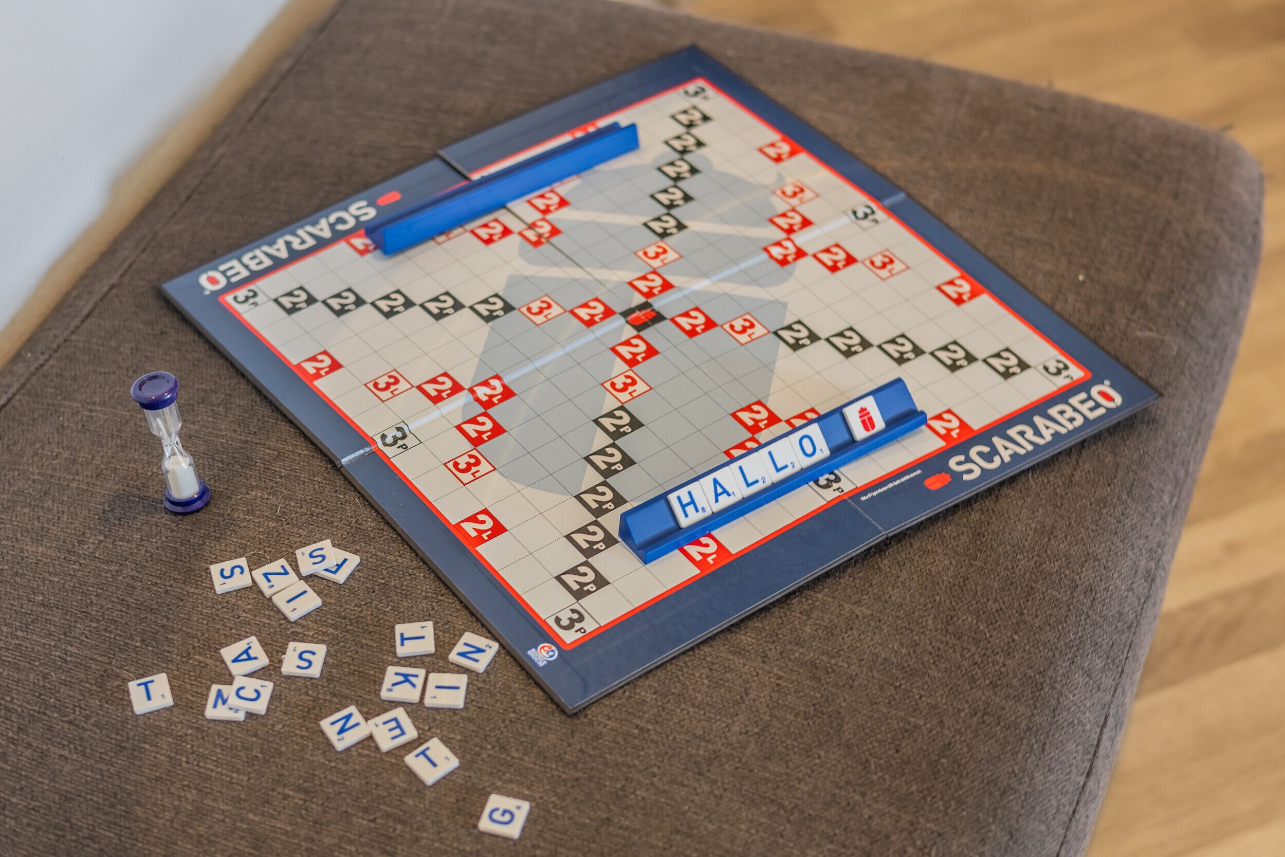 Ein Scrabble-Brettspiel auf einer braunen Oberfläche mit verstreuten Buchstabensteinen und einem Timer.