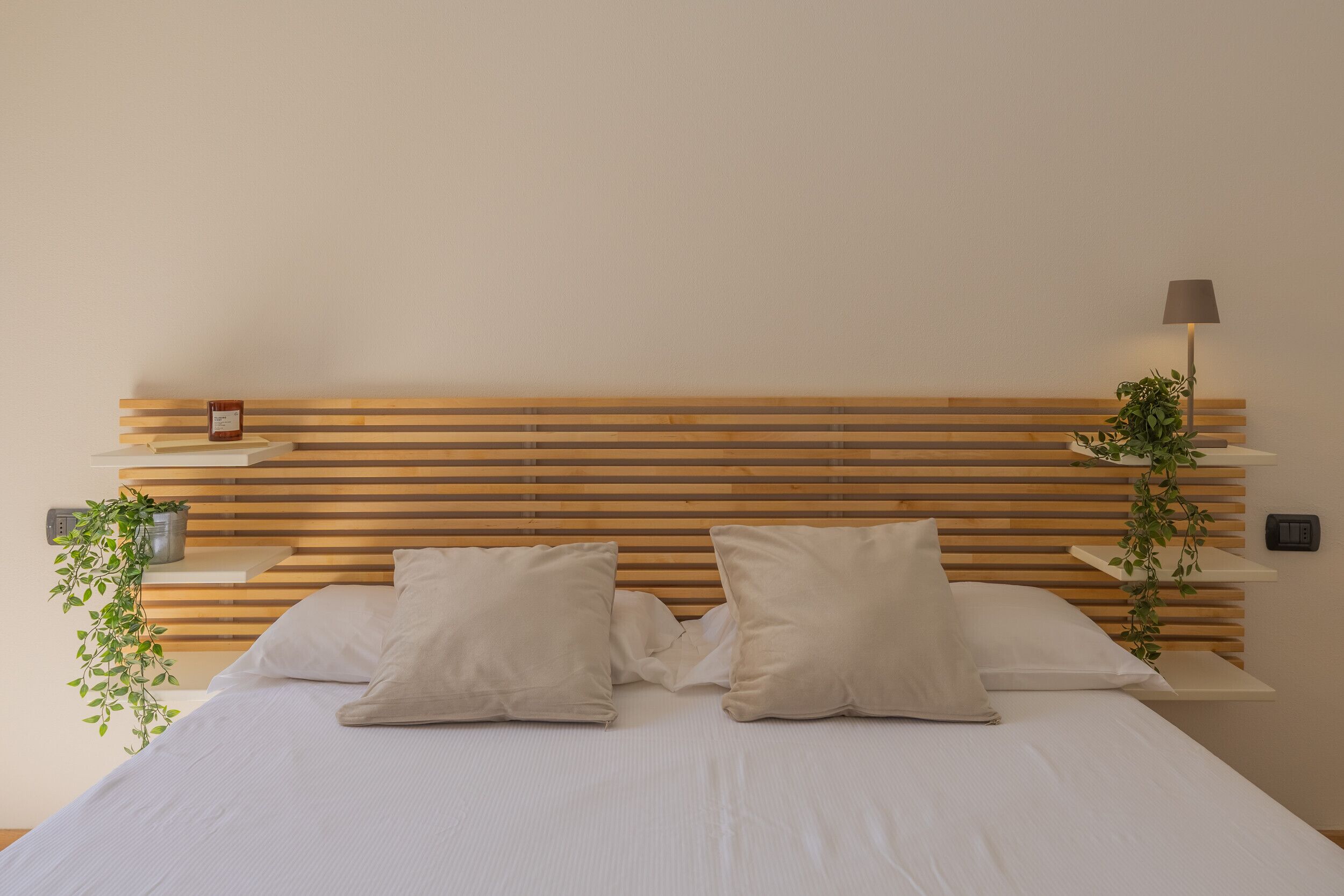 A neatly made bed with two pillows, a wooden slatted headboard, a bedside table with a lamp, and a small plant.