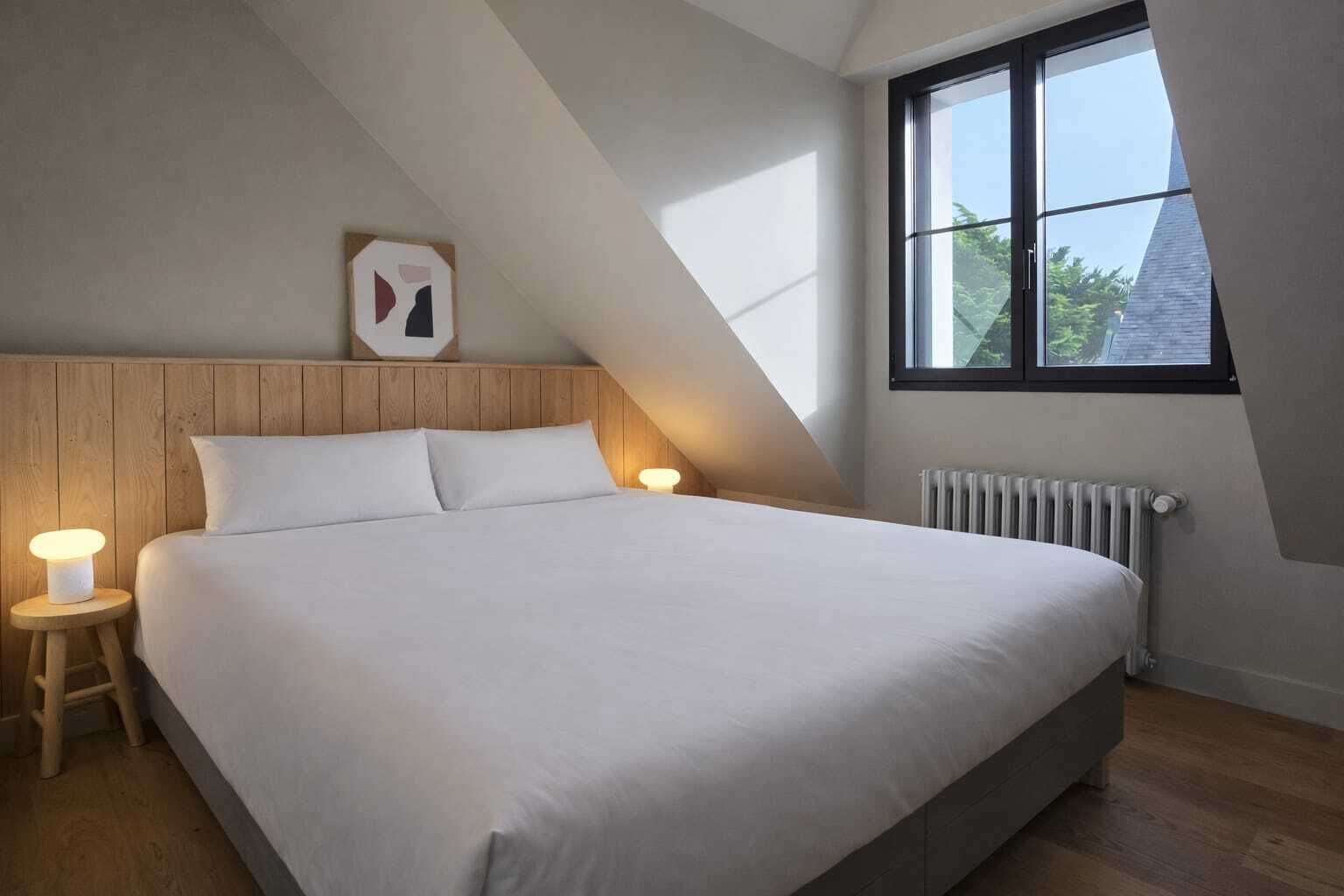 Ett sovrum med ett sluttande tak, en stor säng, två sänglampor, ett element och ett fönster med utsikt över träd.
