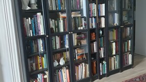 A room with a large bookshelf filled with books, a patterned rug, and a decorative hat on the wall. - Chambre Lavande aux Quatre Vents with Shared Terrace and Garden (Francescas)