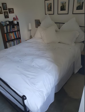 Uma cama bem arrumada com roupa de cama branca, uma estante com livros e quadros nas paredes.