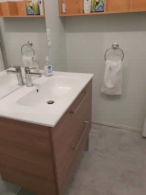 A bathroom with a white sink, two faucets, a wooden cabinet, a towel rack, and a shelf with toiletries.