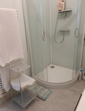 Casa de banho moderna com chuveiro envolto em vidro, um porta-toalhas branco, uma prateleira com artigos de higiene e um pequeno banquinho.