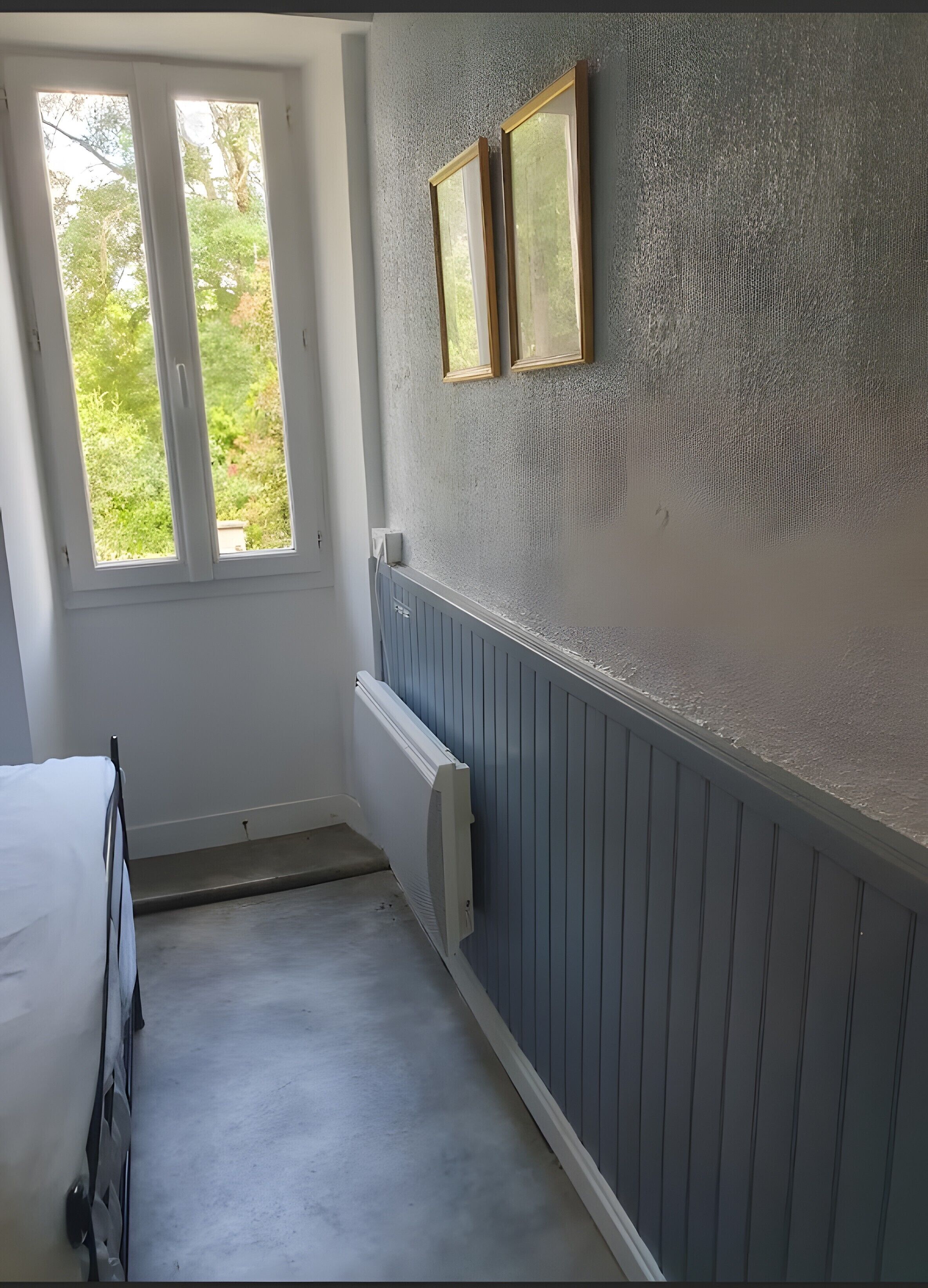 Een kamer met een bed, een radiator, een raam met gordijnen, en twee ingelijste foto’s aan de muur.