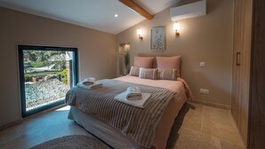 Quarto com uma janela grande, uma cama com almofadas, uma mesa de cabeceira e ar condicionado fixado na parede.