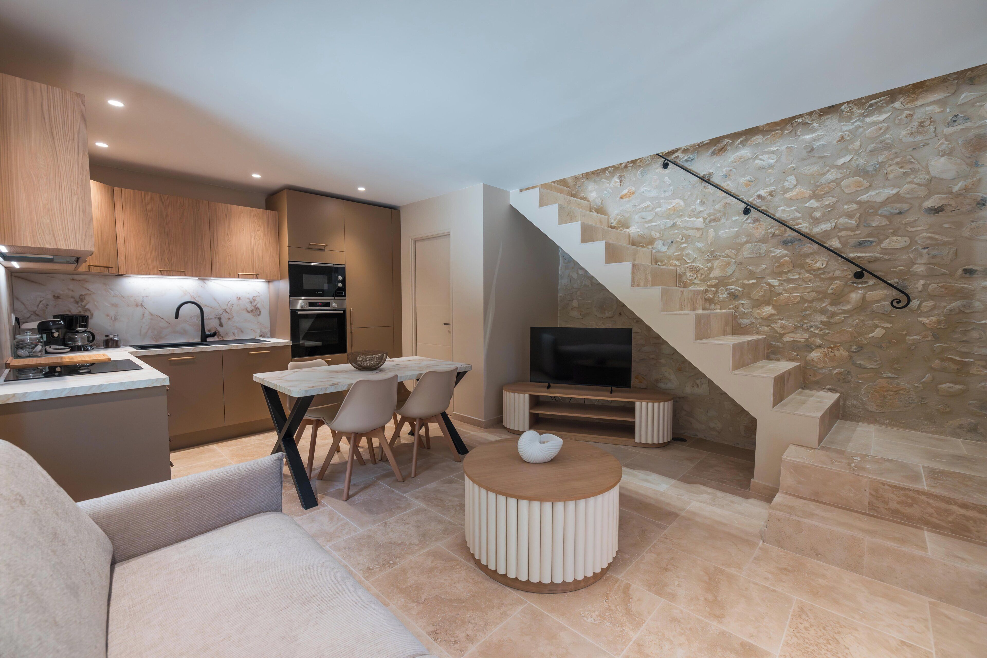 A modern kitchen with wooden cabinets, a dining area with a round table and chairs, a TV, and a stone staircase.