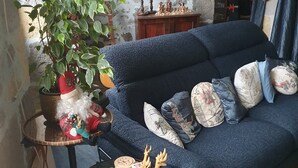 A cozy living room with a blue sofa, a small round table with a plant, and a decorative Santa figure. - Holiday Home Les Quatre Vents with Shared Garden, Terraces, Wi-Fi, and Air Conditioning (Francescas)