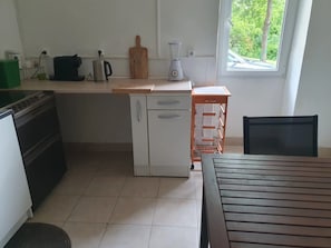 Une cuisine compacte équipée d’une cuisinière, d’un petit réfrigérateur, d’une machine à café, d’un mixeur et d’une fenêtre donnant sur un espace verdoyant.