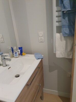 Uma casa de banho com uma pia branca, um espelho, um porta-toalhas com toalhas e produtos de higiene na bancada.