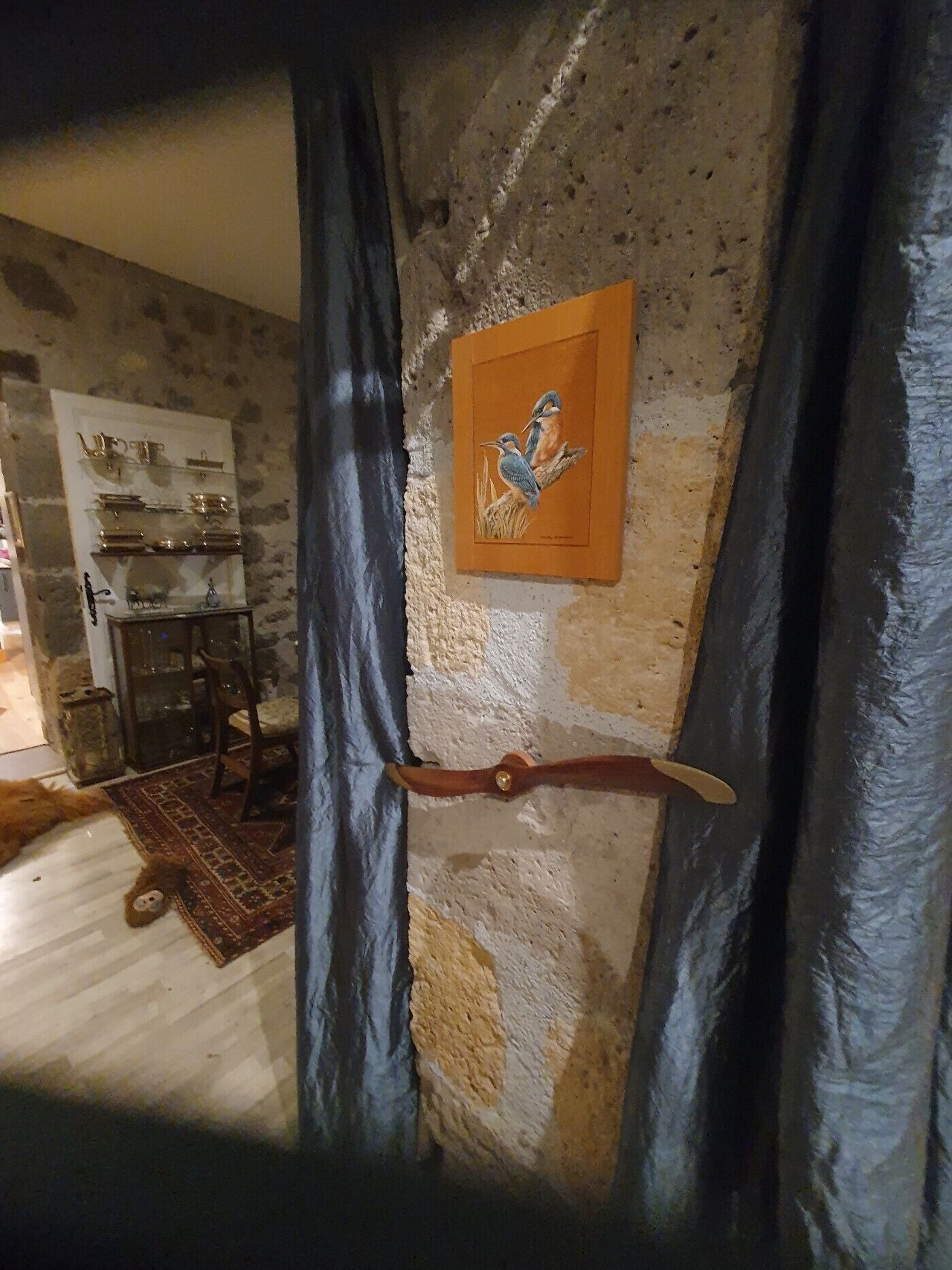 Una habitación con pared de piedra rústica, una obra de arte enmarcada con pájaros, una puerta de madera y una pequeña mesa con objetos encima.