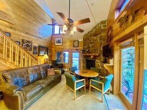 A cozy cabin interior with a leather sofa, wooden dining table, stone fireplace, and a ceiling fan. - Cabin Feel w/ Loft - Amazing Views -Pool.  Near RMNP, Granby Ranch & Winter Park (Granby)