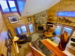 Interior acogedor de una cabaña con chimenea de piedra, paredes de madera, comedor y vistas a través de las ventanas.