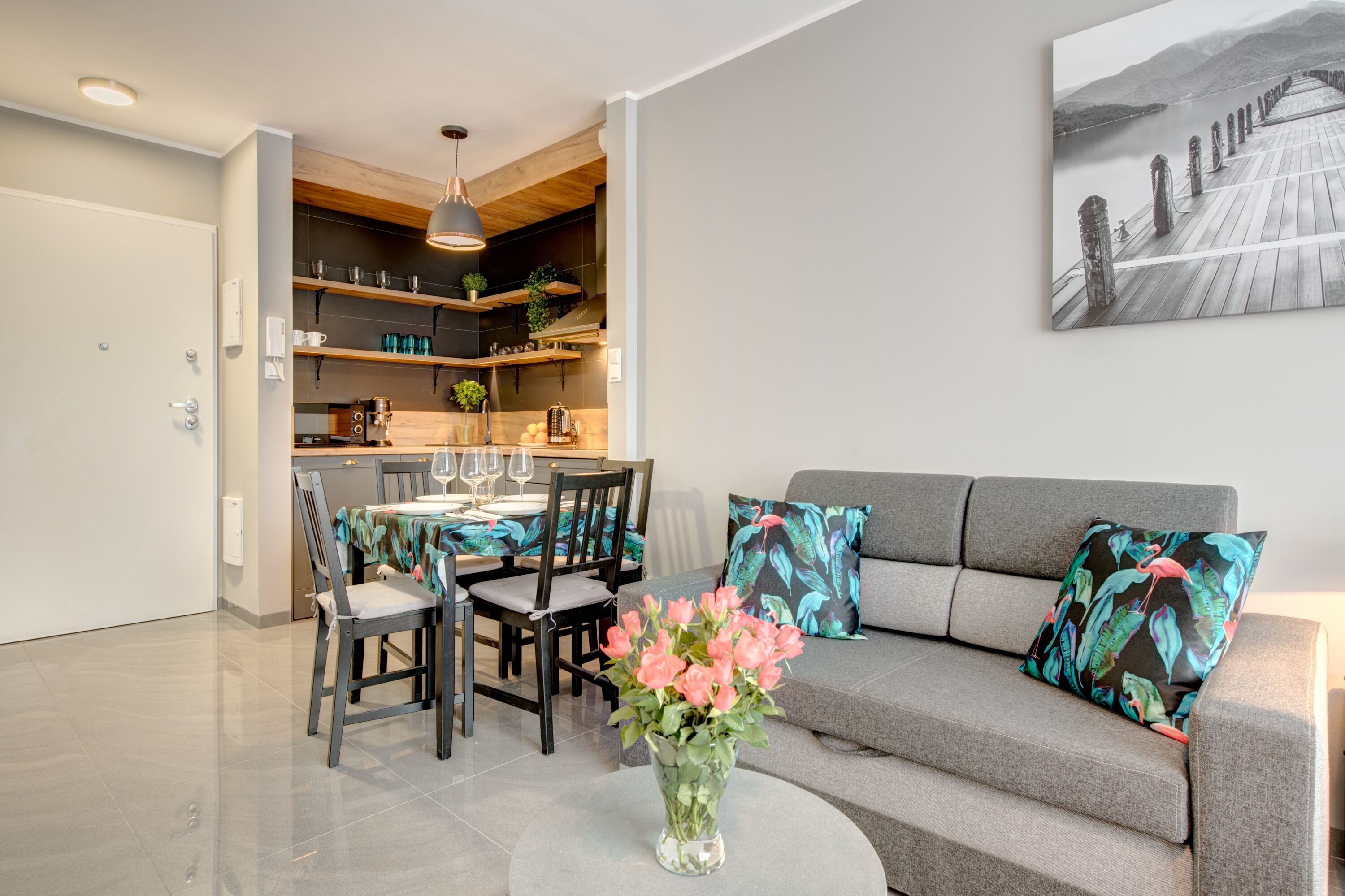 Ein modernes Wohnzimmer mit einem grauen Sofa, einem Esstisch mit Stühlen, einer Blumenvase und einem durchgehenden Türrahmen sichtbaren Küchenbereich.