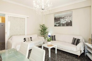 Ein Wohnzimmer mit zwei weißen Sofas, einem Glastisch, einer Chandelüster, einer Topfpflanze und einem gerahmten Stadtbild an der Wand.