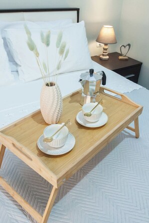 Ein Holztablett mit weißen Geschirr und einer Kaffeekanne auf einem Bett mit weißer Bettwäsche und einem Nachttisch mit Lampe.