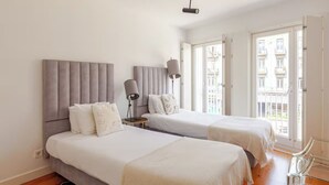 A hotel room with two beds, a bedside table with a lamp, and a view of the city through large windows. - Stylish Retreat with Rare Rooftop Pool | 2BR, 2BA (Lisboa)