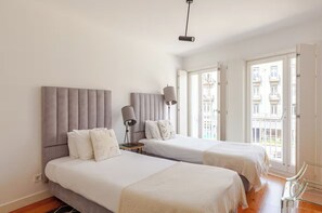 Ein Hotelzimmer mit zwei Betten, einem Nachttisch mit Lampe und Blick auf die Stadt durch große Fenster.