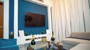 Ein modernes Wohnzimmer mit blauer Akzentwand, einem Flachbildfernseher über einem Glastisch und einem beigefarbenen Sofa.