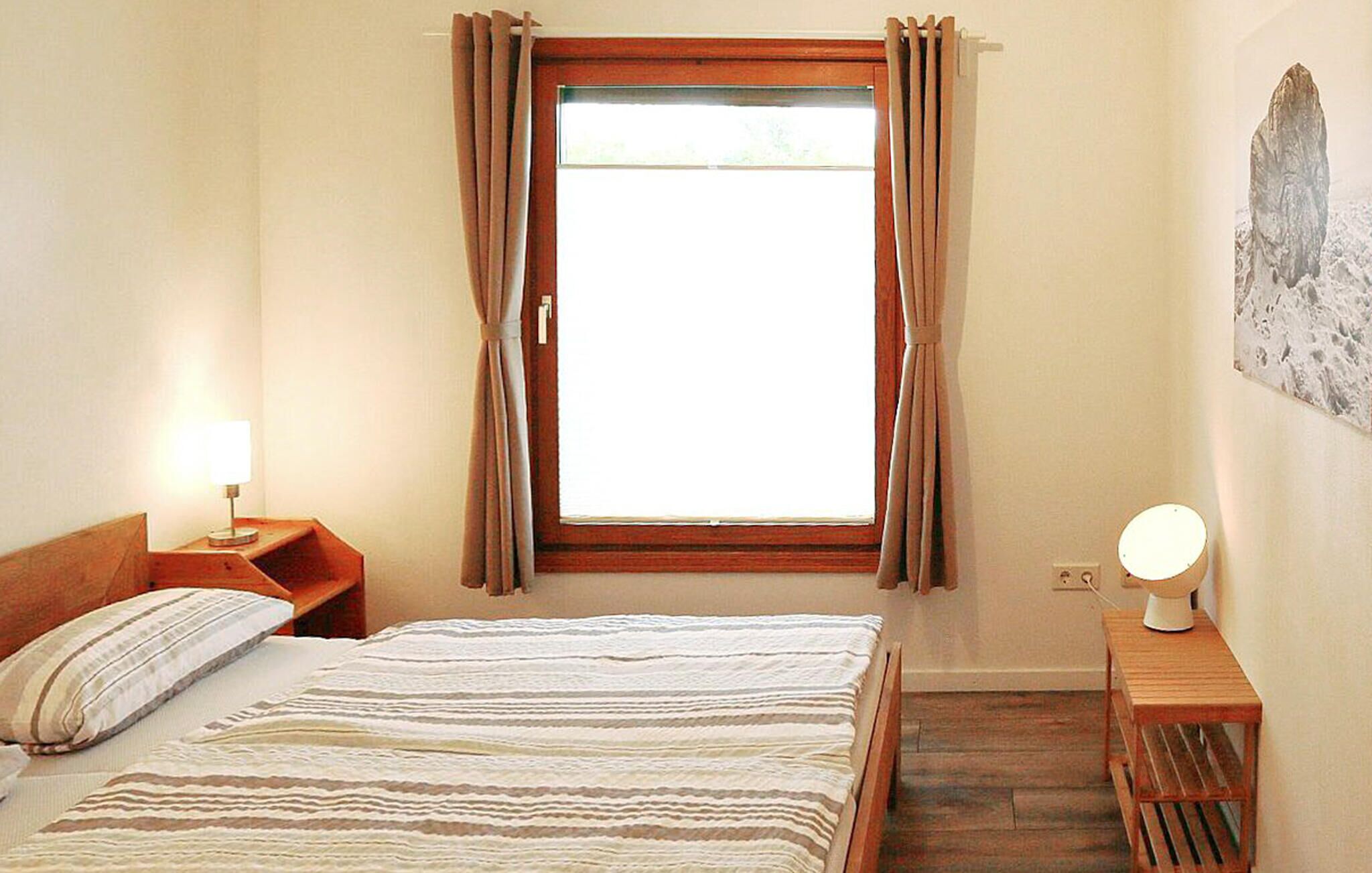 A bedroom with a bed, a nightstand with a lamp, a window with curtains, and a wall-mounted lamp.