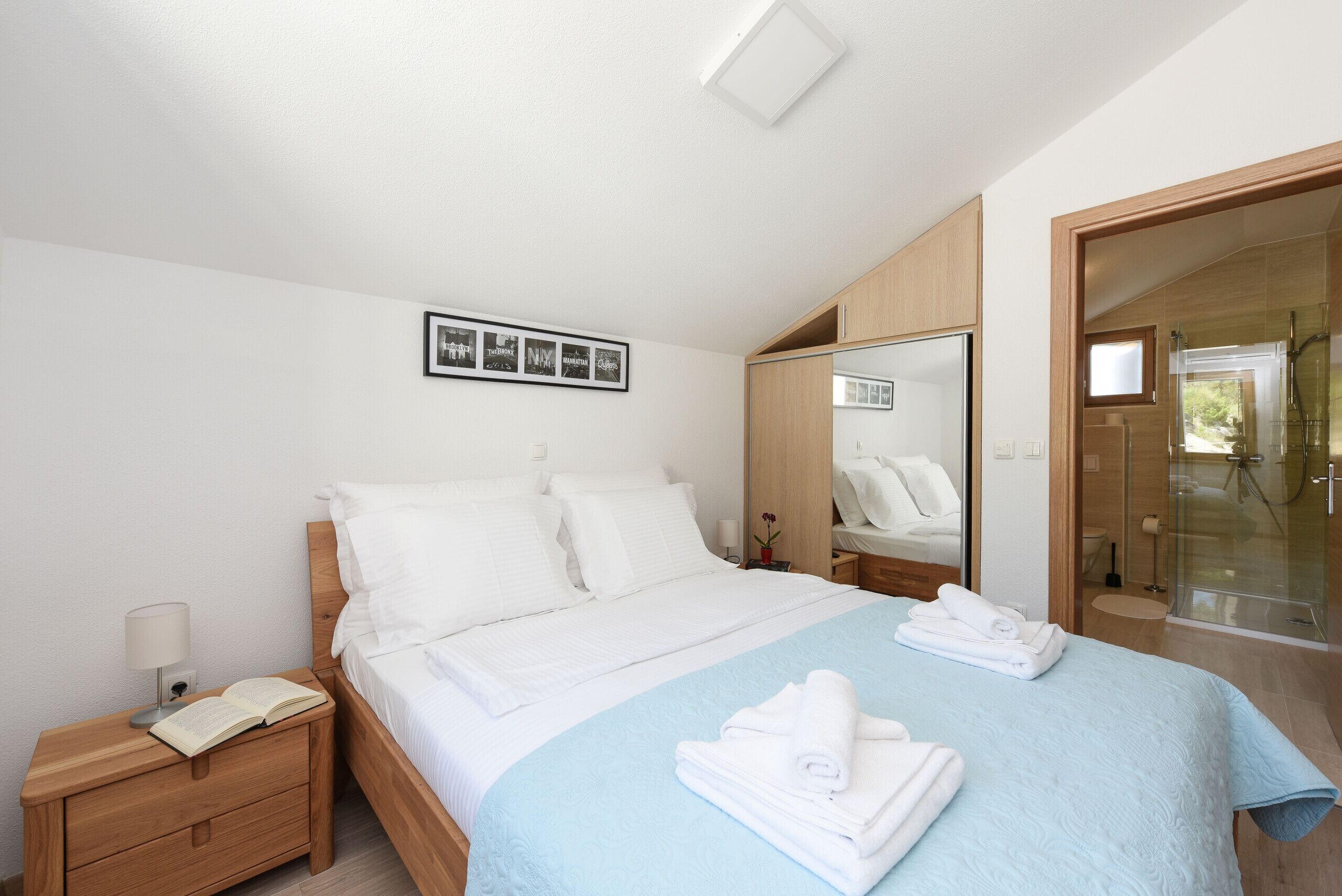 Una habitación de hotel moderna con una cama bien hecha, una mesita de noche con lámpara, un espejo y una zona de ducha.