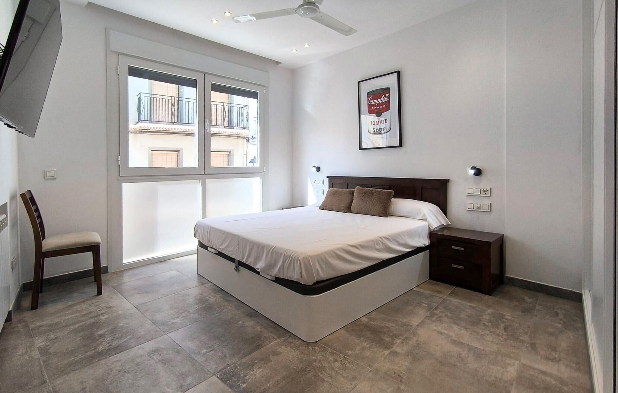 A bedroom with a bed, a chair, a nightstand, a framed picture, and a window.