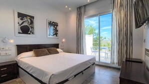 Ein modernes Schlafzimmer mit einem großen Bett, zwei an der Wand befestigten Kunstwerken, einer Schiebeglastür zu einem Balkon und einem dunklen Nachttisch aus Holz.