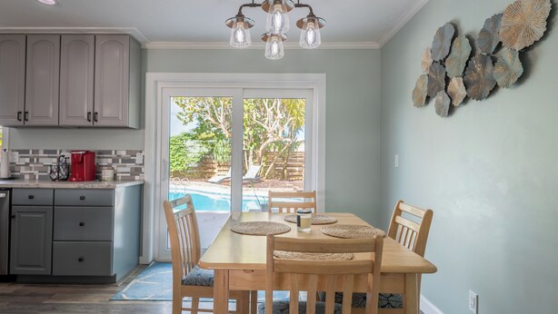 A kitchen with grey cabinets, a dining table with chairs, a red coffee maker, and a view of a backyard with a pool. - Space Coast Retreat With Heated Pool (Merritt Island)