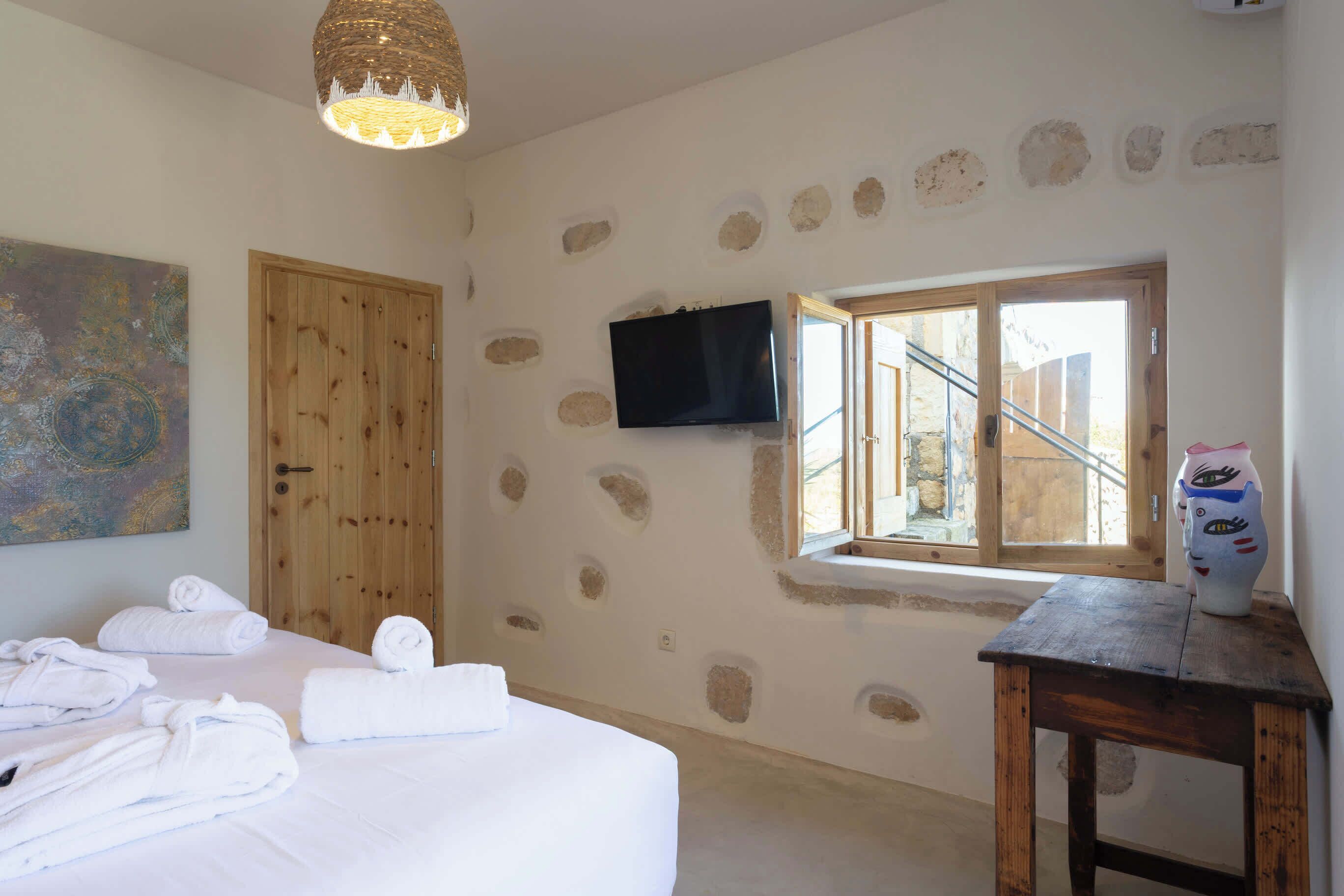 A bedroom with a bed, towels, a television, a window, and a stone wall.