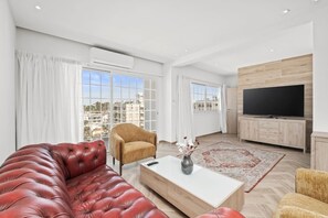 Ein modernes Wohnzimmer mit einer Ledersofa, einem Holztisch, einem Fernseher und einem großen Fenster mit Vorhängen.