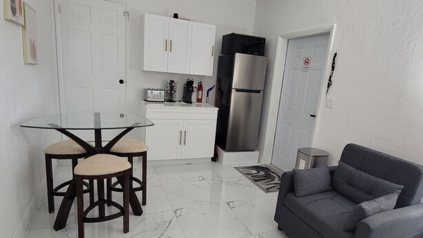 A compact kitchen with white cabinets, a stainless steel refrigerator, a glass-top table with two chairs, and a grey sofa in a living area.