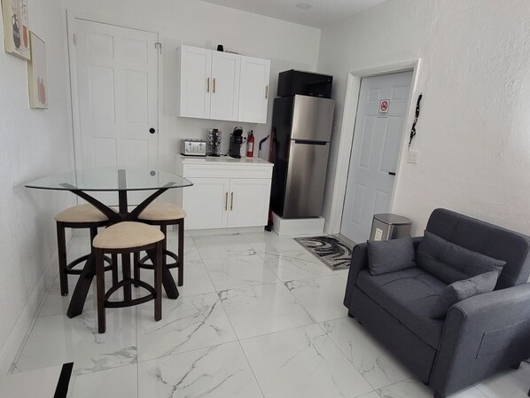 Cocina compacta con armarios blancos, un refrigerador de acero inoxidable, una mesa de vidrio con dos sillas y un sofá gris en la sala.