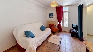 Ein Zimmer mit weißem Sofa, einem kleinen Tisch, einem Sessel, einem Fernseher und einem Fenster mit Vorhängen.