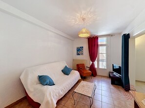 Ein Zimmer mit weißem Sofa, einem kleinen Tisch, einem Sessel, einem Fernseher und einem Fenster mit Vorhängen.