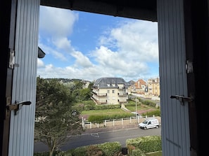 Blick durch ein Fenster auf ein Gebäude, im Hintergrund ein großes Haus, ein Auto auf der Straße und viel Grün.