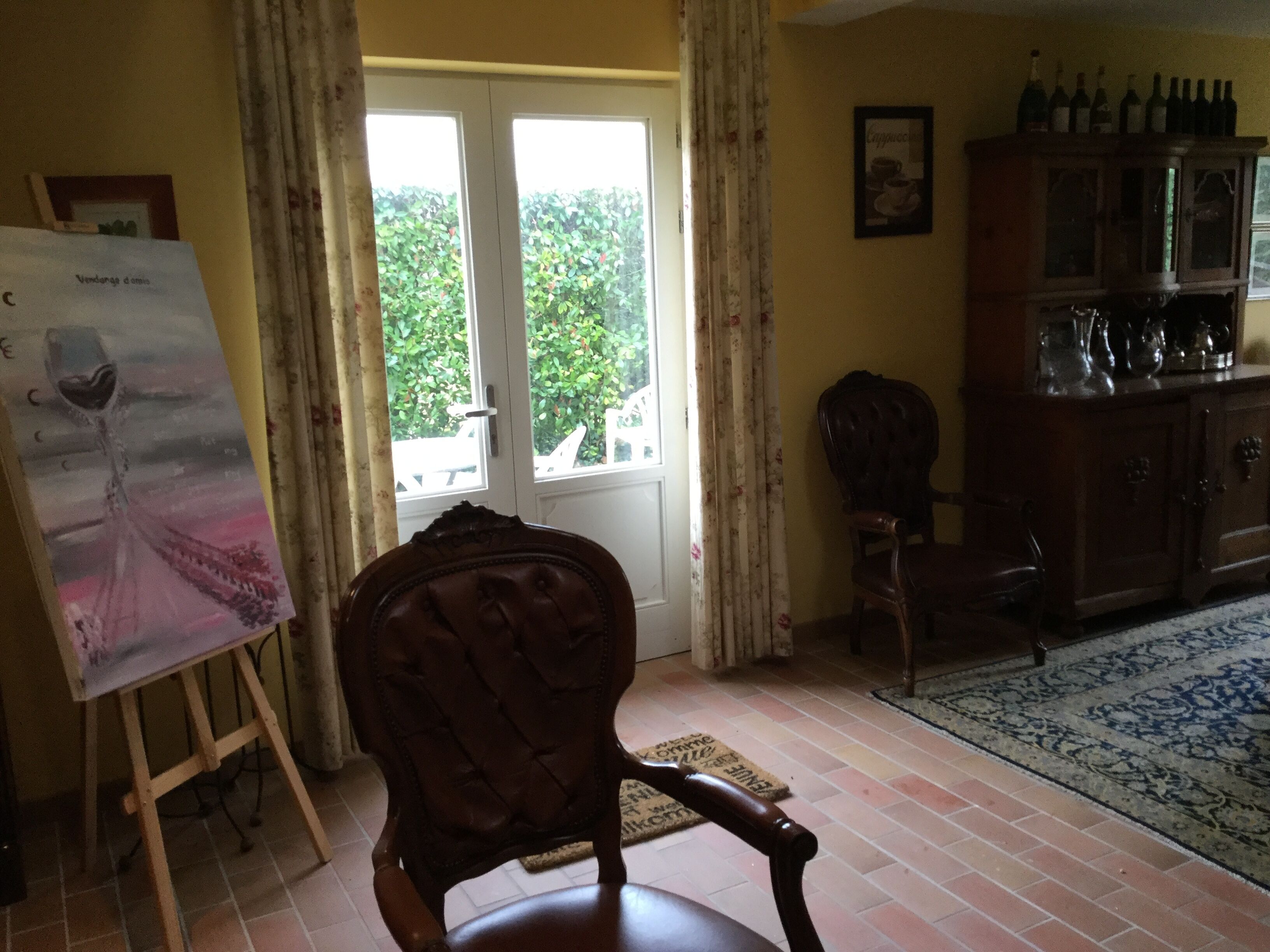 Una habitación con una silla de madera, un cuadro sobre caballete, un aparador con botellas y vistas por una ventana.