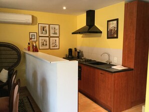 Eine kompakte Küche mit weißer Arbeitsplatte, einem schwarzen Spülbecken und Holzschränken. An der Wand befinden sich gerahmte Bilder und eine schwarze Dunstabzugshaube über dem Herd.
