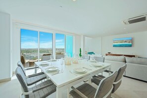 Un comedor moderno con una mesa blanca para cuatro, ventanas amplias con vistas a la ciudad y un sofá gris al fondo.