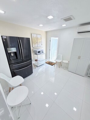 Una cocina moderna con un refrigerador negro, sillas blancas, un microondas y una cafetera sobre un mueble blanco.