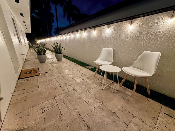 Un pasillo con un felpudo, plantas en macetas y dos sillas blancas frente a una pared con textura.