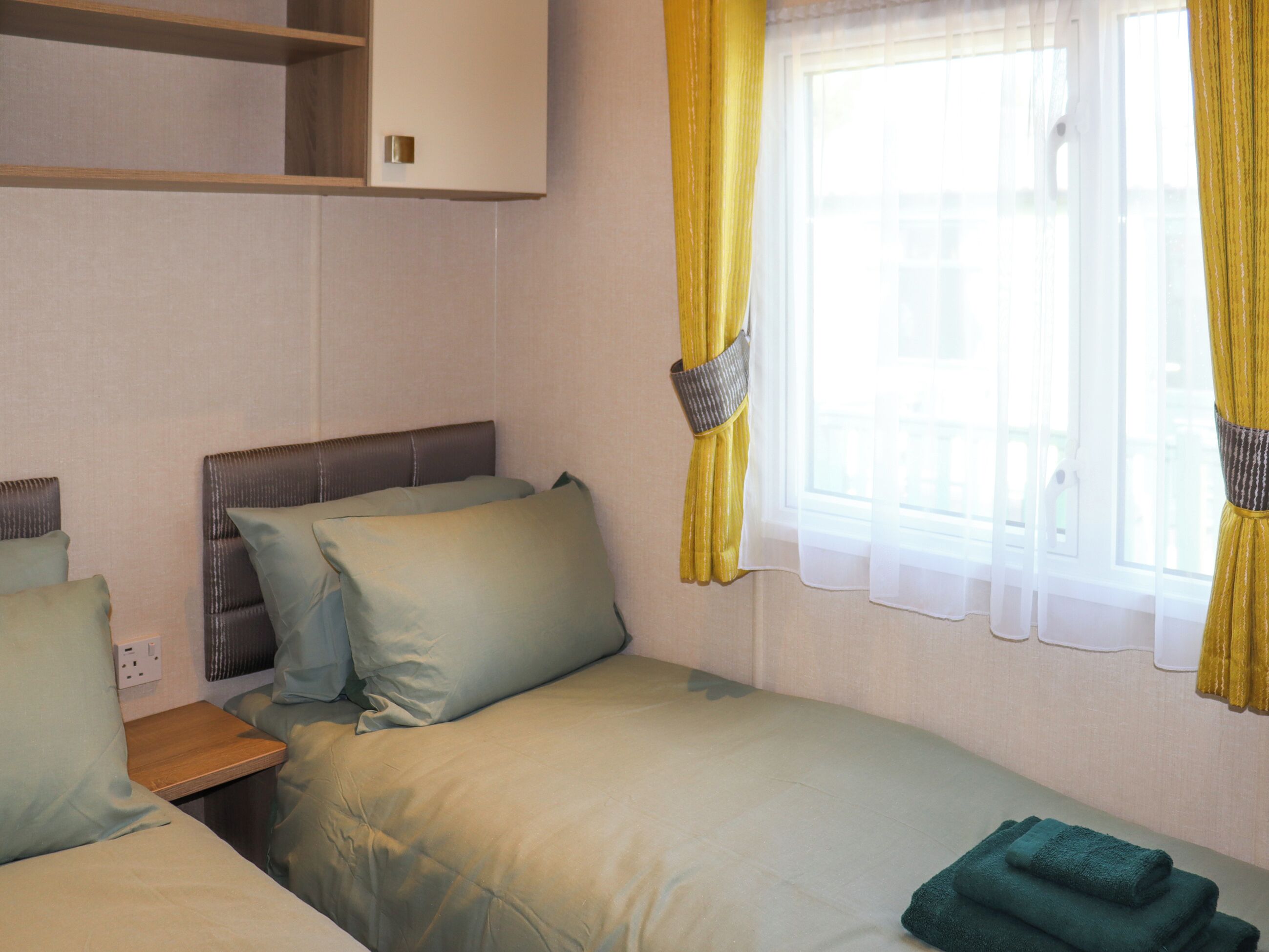 A single bed with a headboard, a bedside table, and a window with yellow curtains.