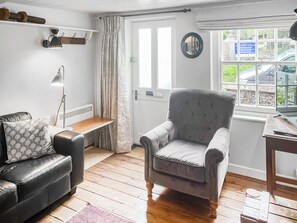 Ein Zimmer mit grauem Sessel, einer schwarzen Ledersofa, einem kleinen Holztisch, einer Stehlampe, einem Fenster mit Blick auf Autos und Grünflächen und einer Tür mit Spiegel.