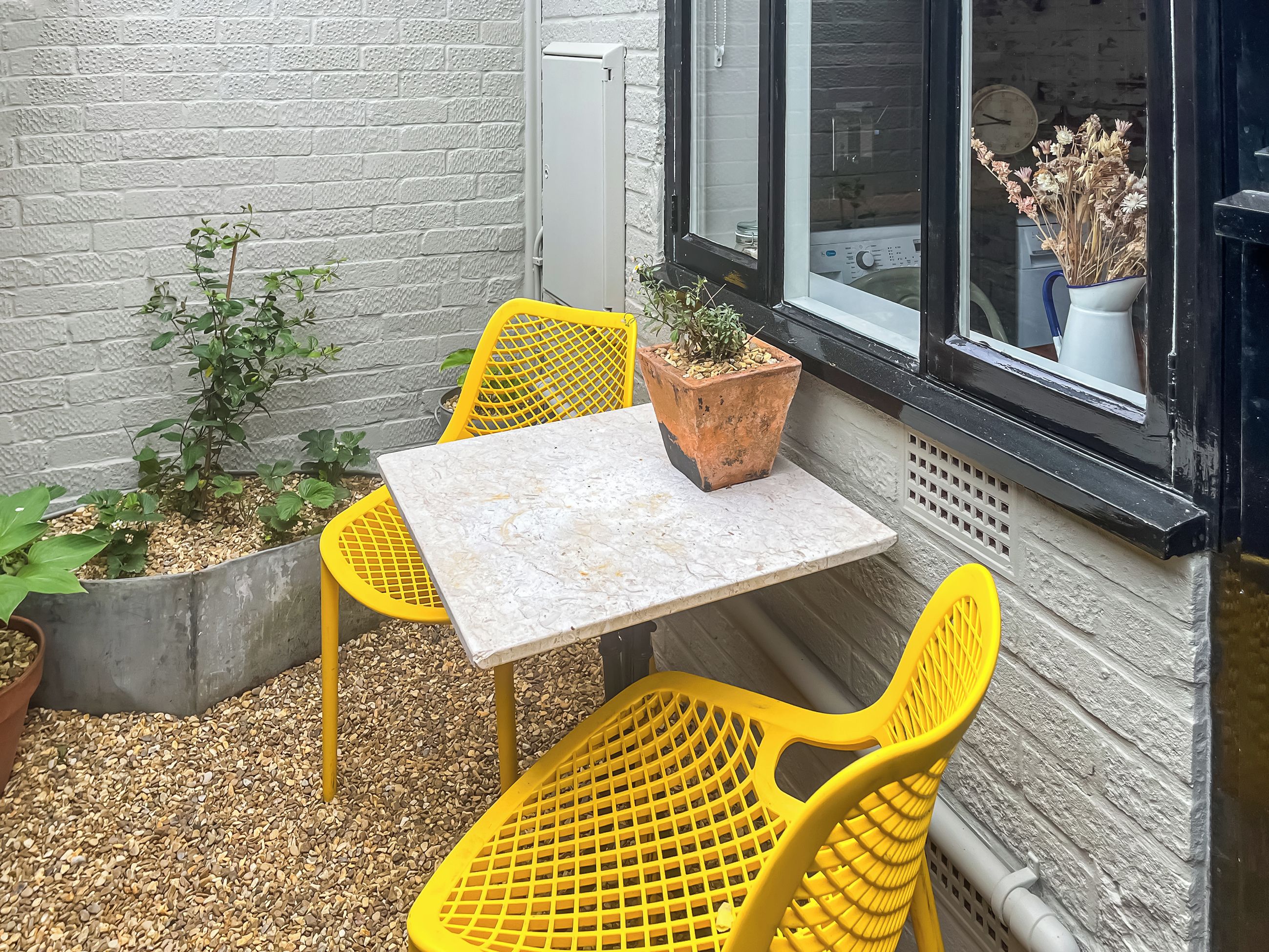 A small outdoor seating area with a table and two chairs, a potted plant, and a window with a view of the street.