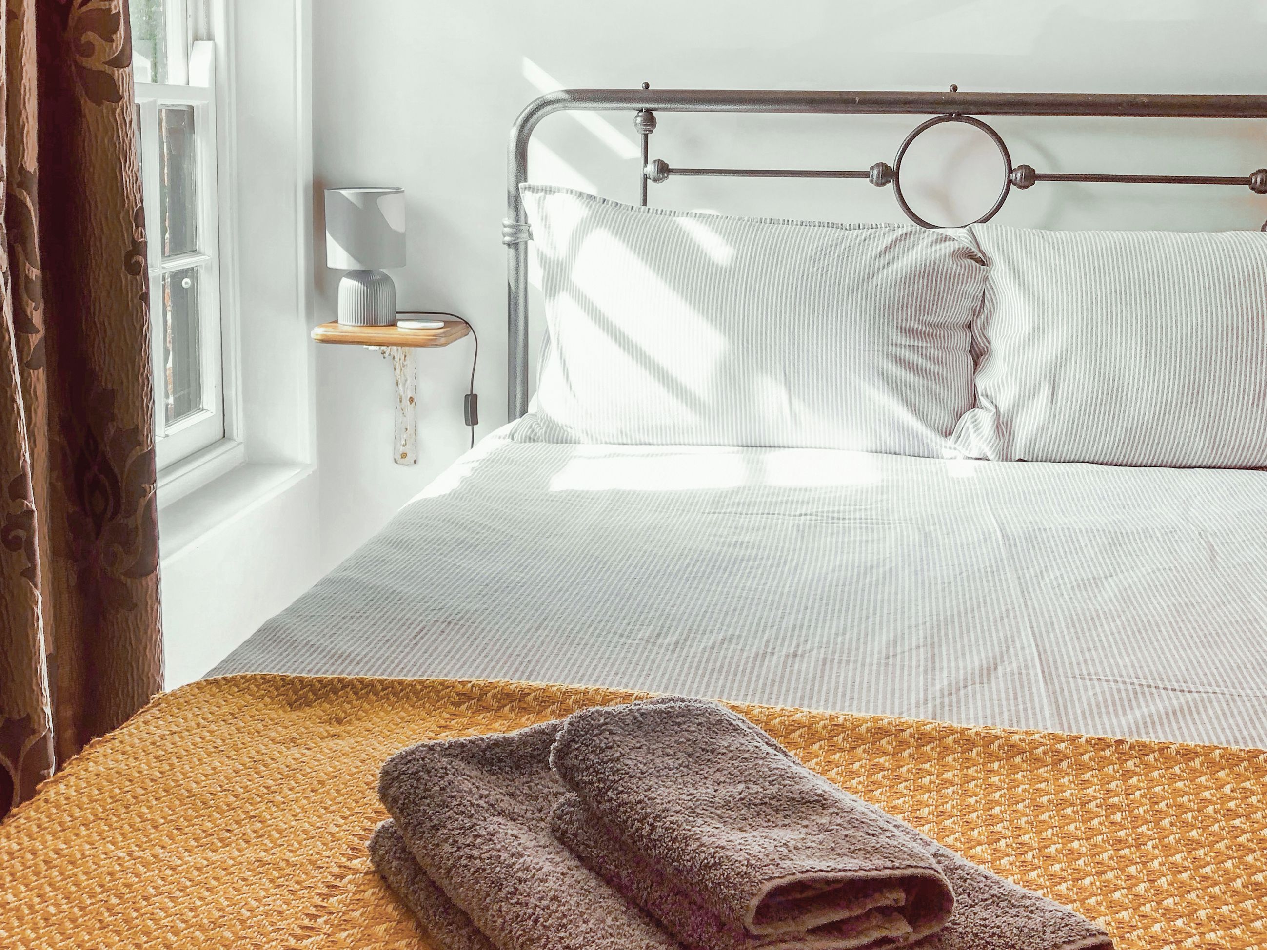 A neatly made bed with a striped pillow and a rolled towel on a woven mat.