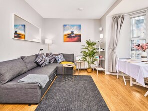 Ein modernes Wohnzimmer mit einem grauen Sofa, einem kleinen Tisch, einem Blumentopf, einer weißen Regaleinheit und Kunstwerken an den Wänden.
