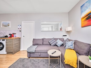 Ein modernes Wohnzimmer mit einem grauen Sofa, einem kleinen runden Tisch, einer Waschmaschine und einer Küchenzeile mit Spüle und Arbeitsplatte.