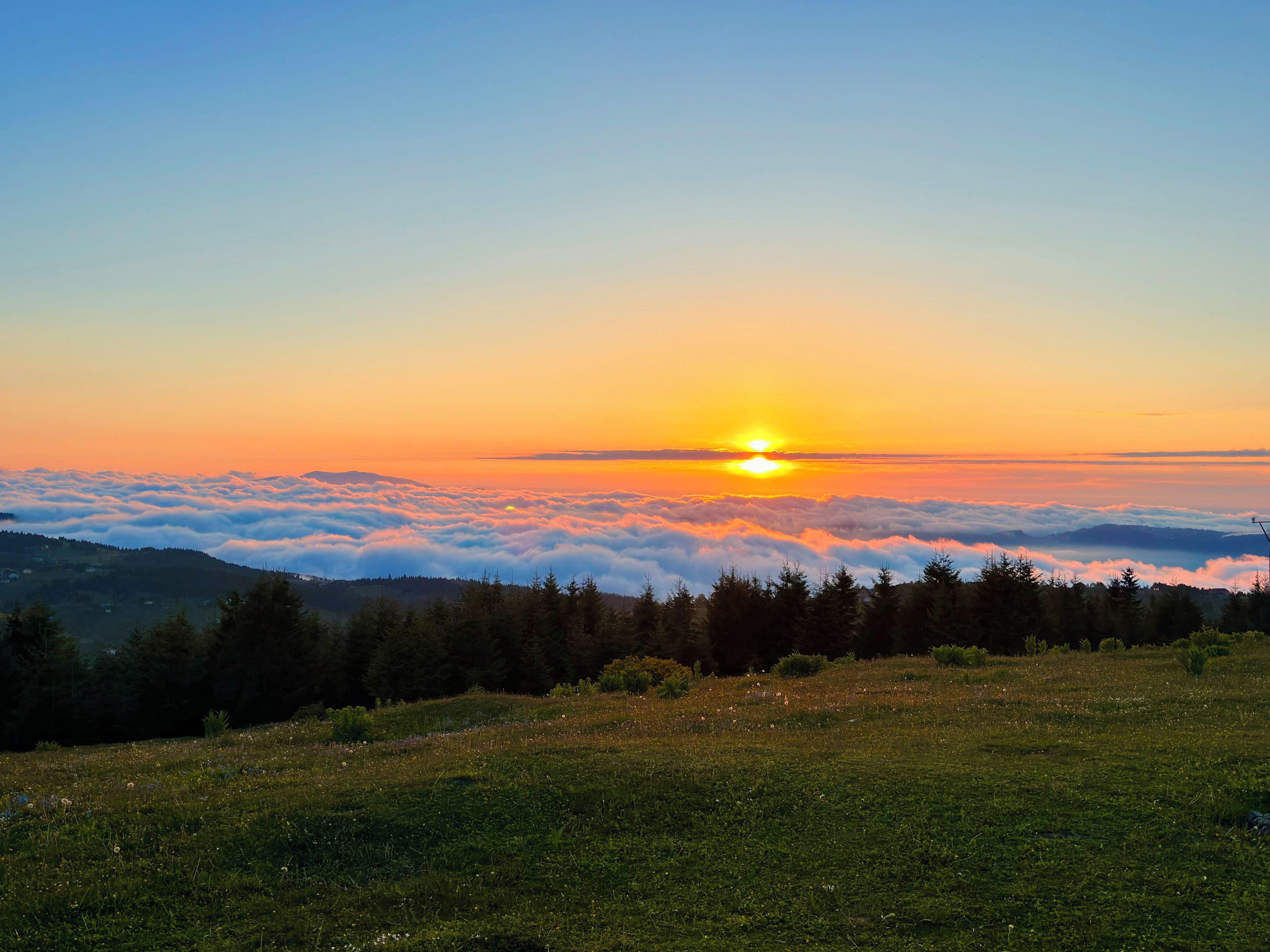 Un lever de soleil sur une chaîne de montagnes boisées, avec une vue sur le soleil au-dessus des nuages.
