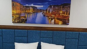 Habitación de hotel con una cama, dos almohadas, un cabecero azul y un cuadro de un paisaje urbano.