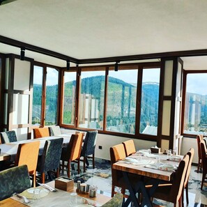 Comedor con grandes ventanales que ofrecen vistas a la montaña, mesas y sillas de madera, y piso de baldosas.