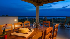 Ein Holztisch mit Käselaib, Weingläsern und einer Flasche, vor einer Blick auf das Meer im Abenddämmerung.
