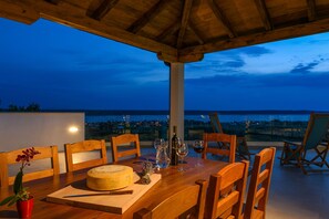 Ein Holztisch mit Käselaib, Weingläsern und einer Flasche, vor einer Blick auf das Meer im Abenddämmerung.